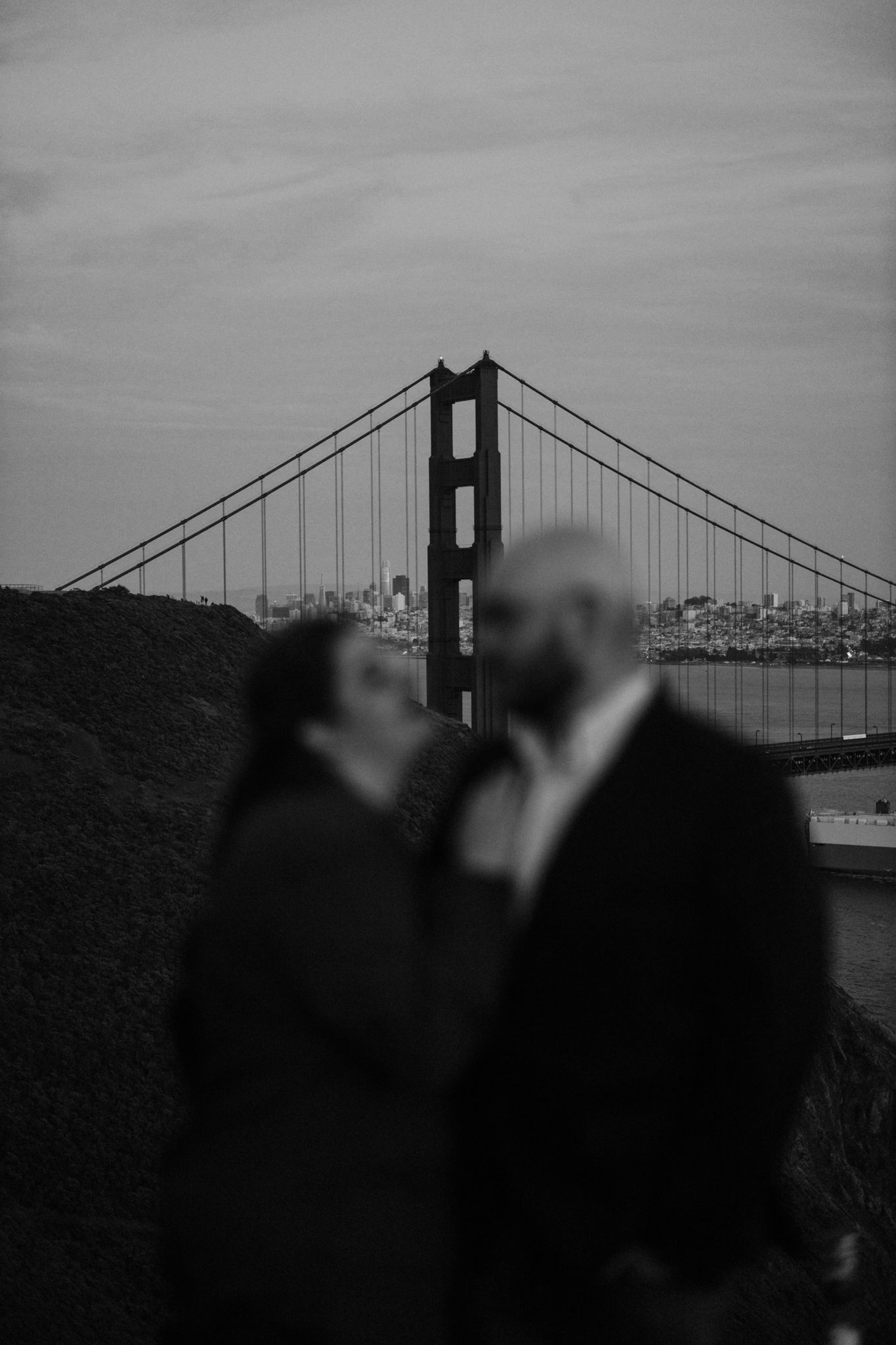 Black and white photo with couple blurred and the golden gate bridge in focus in the background near San Francisco, California