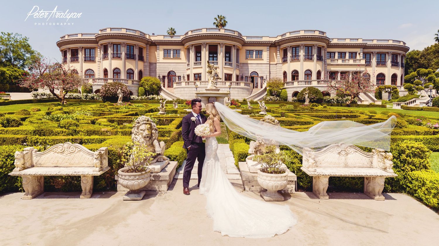 Elegant wedding photo at grand estate with manicured gardens and stone benches framing the path.