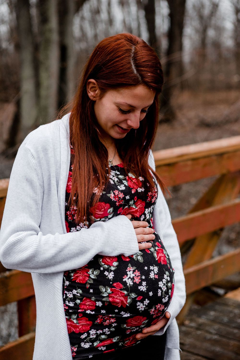 Expectant parent in white cardigan and floral dress lovingly touches baby bump in autumn forest.
