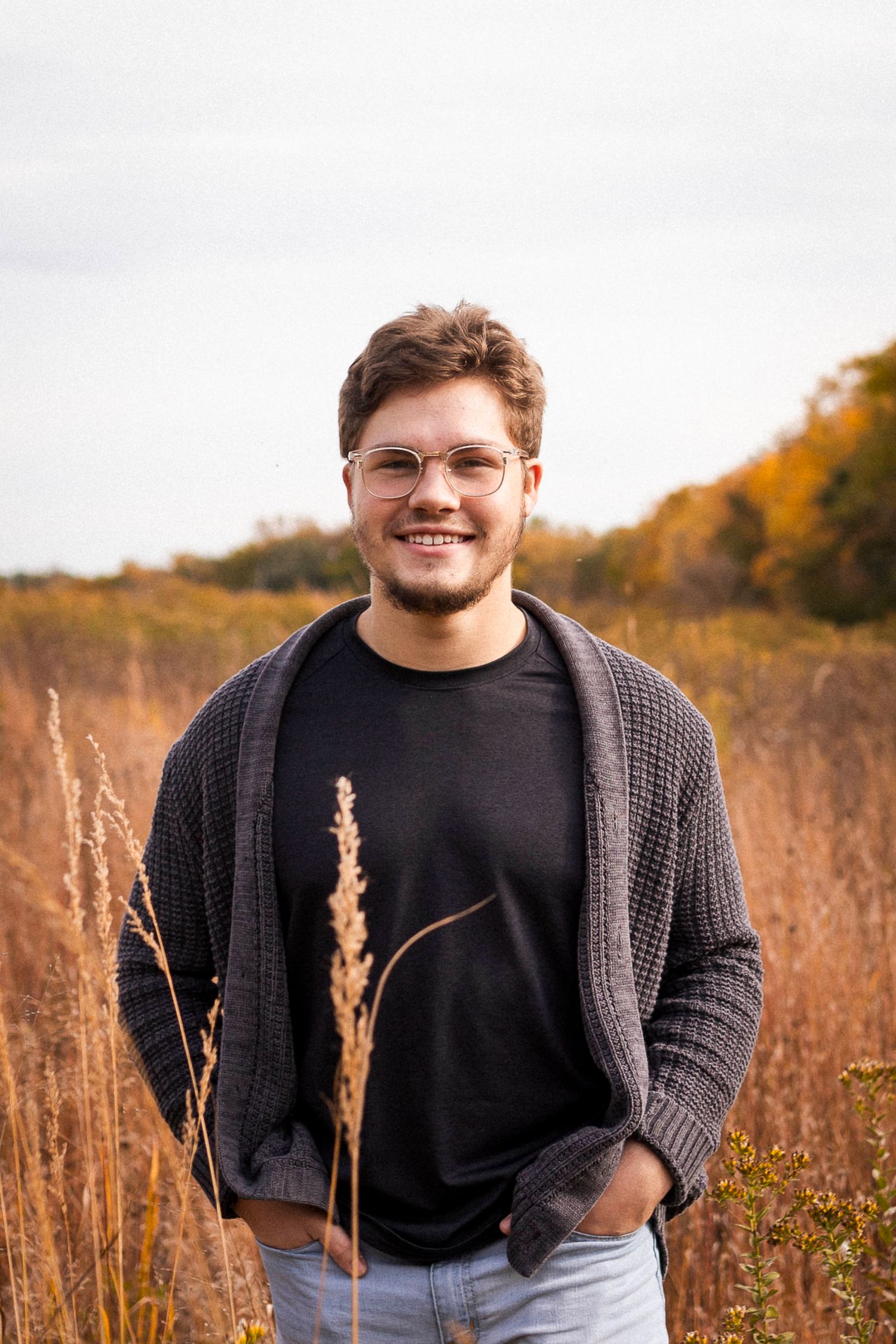 Senior Portrait. Boy standing in golden field wearing black shirt and grey sweater. Smiling with glasses and short hair