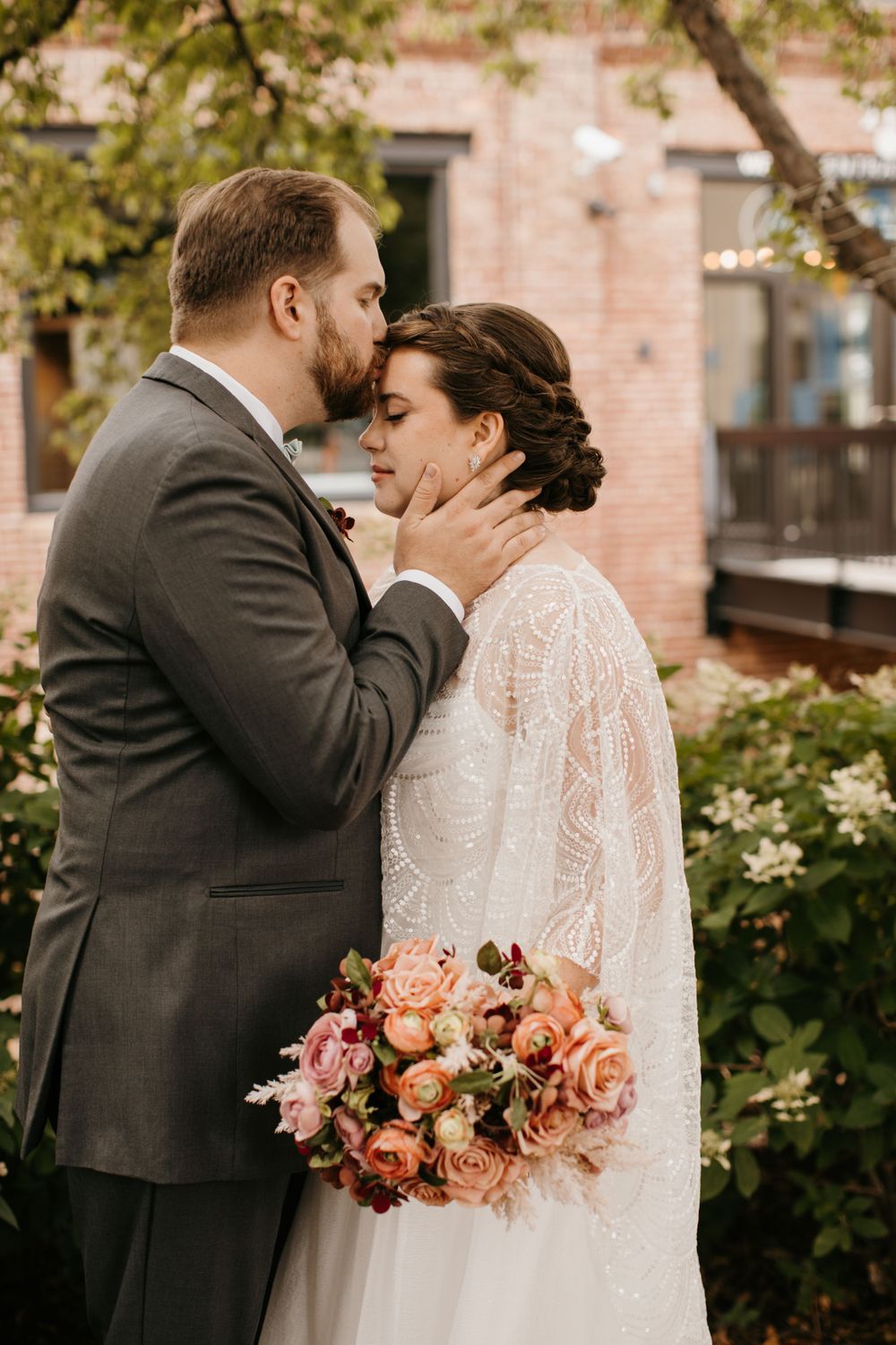 Groom tenderly kisses bride on forehead while cradling her face, bride holds colorful bouquet of pink and coral roses.