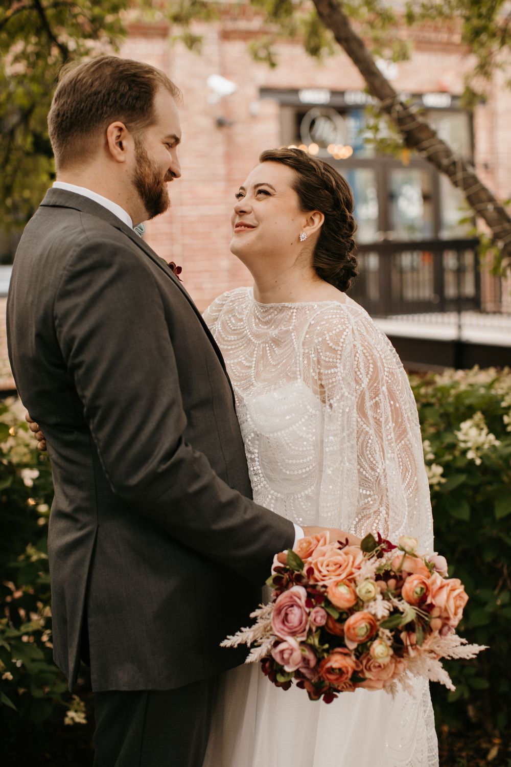 Bride in lace cape gown smiles lovingly at groom while holding a pink floral bouquet.