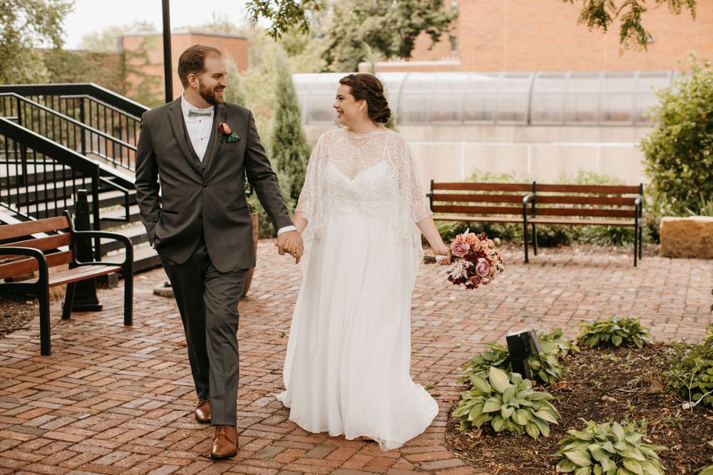 Bride and groom walk hand in hand on brick pathway, bride carries colorful bouquet in flowing white wedding dress.