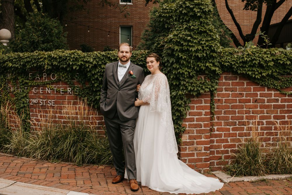 Bride and groom pose together in front of ivy-covered brick wall, bride wears lace cape wedding dress.