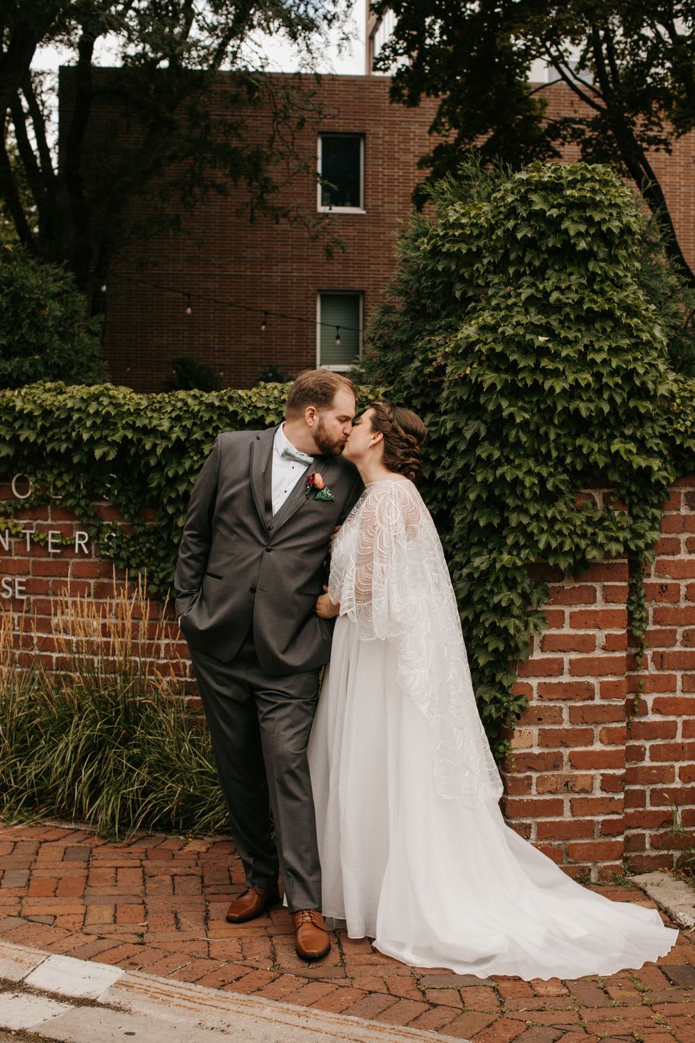 Bride and groom share a romantic kiss on brick sidewalk in front of ivy-covered wall outdoors.