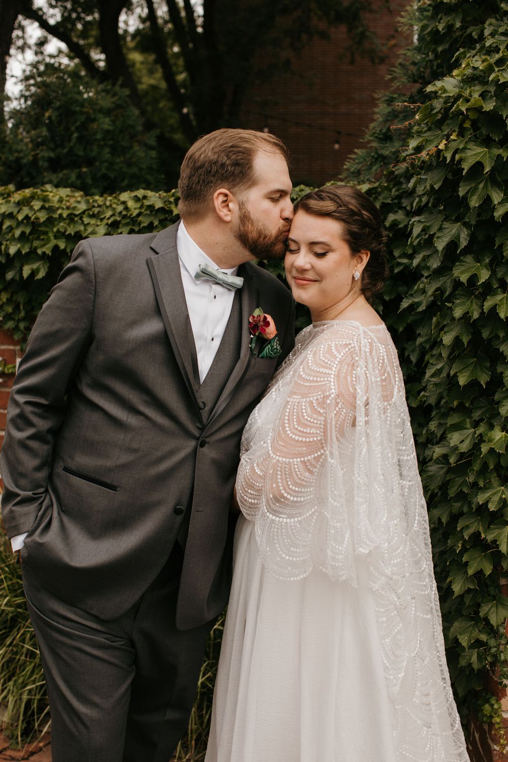 Groom kisses bride on temple as she leans into him, bride wears beaded lace cape wedding gown.