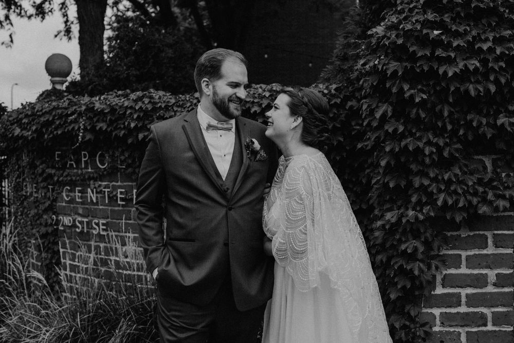 Black and white photo of bride and groom smiling at each other in front of ivy-covered brick wall.