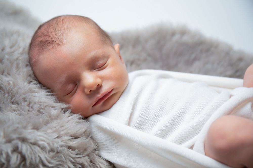 Sleeping newborn on fluffy gray blanket wearing white clothing.