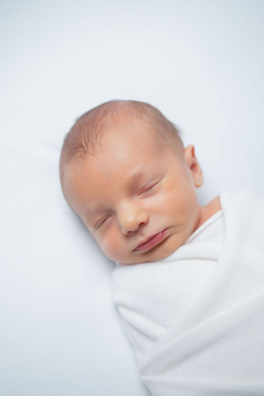Peaceful sleeping infant wrapped in white swaddling against light backdrop.