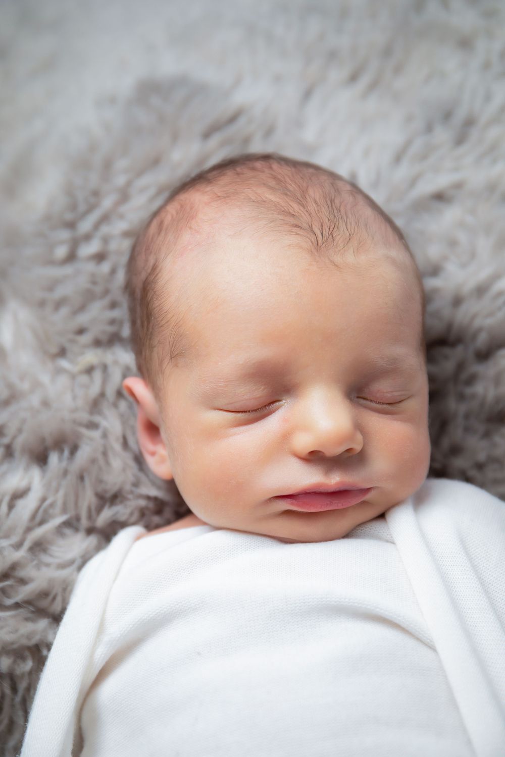 Gentle portrait of sleeping newborn swaddled in white on gray fur backdrop.
