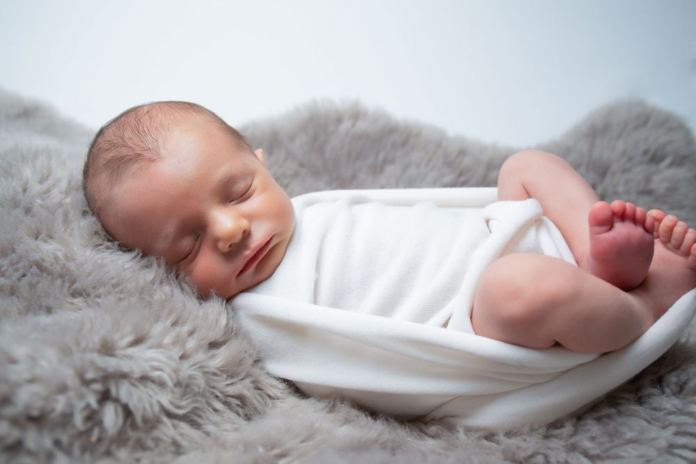 Peaceful newborn baby sleeping on soft gray fur wearing white clothing.