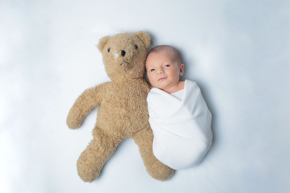 Tiny baby wrapped in white blanket lies beside soft brown teddy bear on light backdrop.