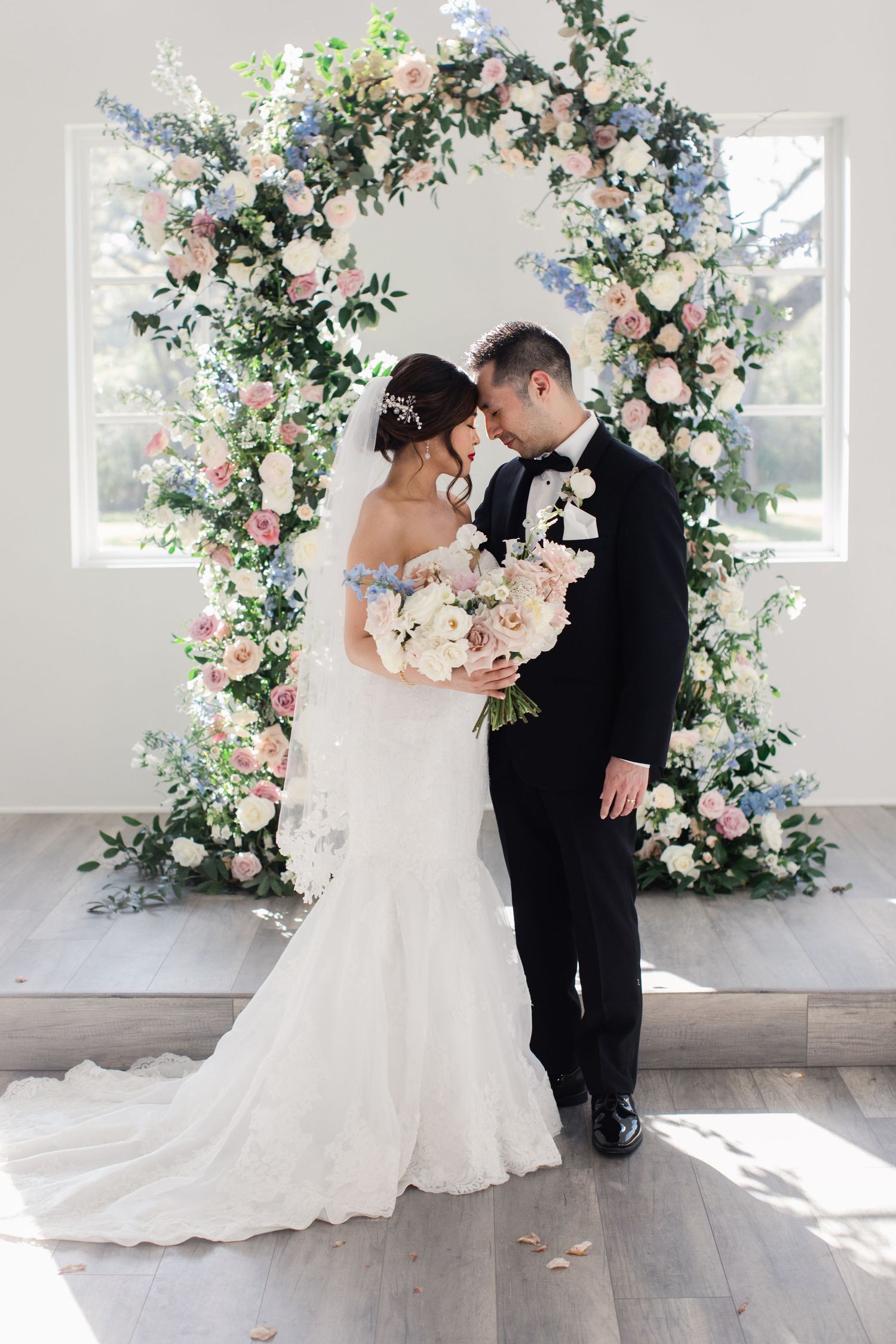 A wedding couple poses under a stunning circular floral arch with pink and white blooms against a bright window.