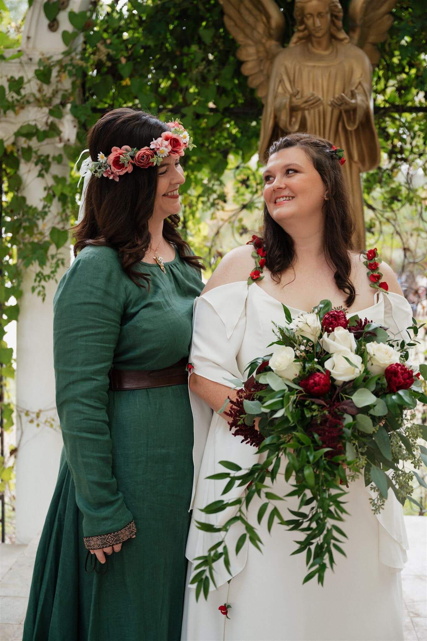 Two attendants share a moment under greenery, one wearing emerald dress and one in white with floral bouquet.