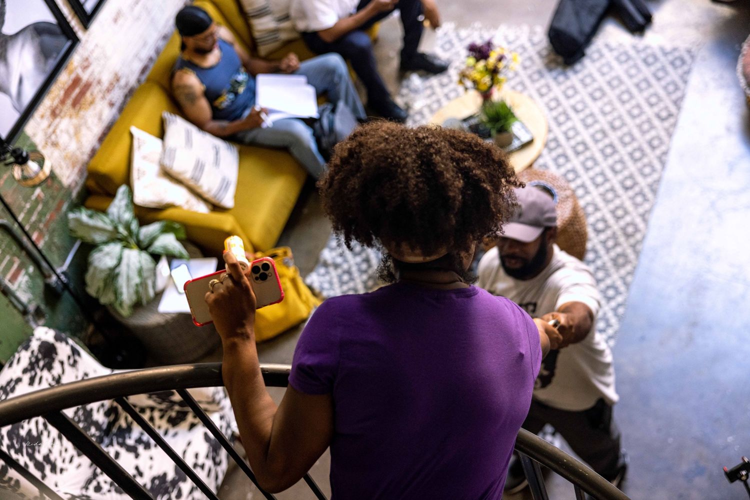 People gather around a coffee table covered in books and papers in a brightly lit room.