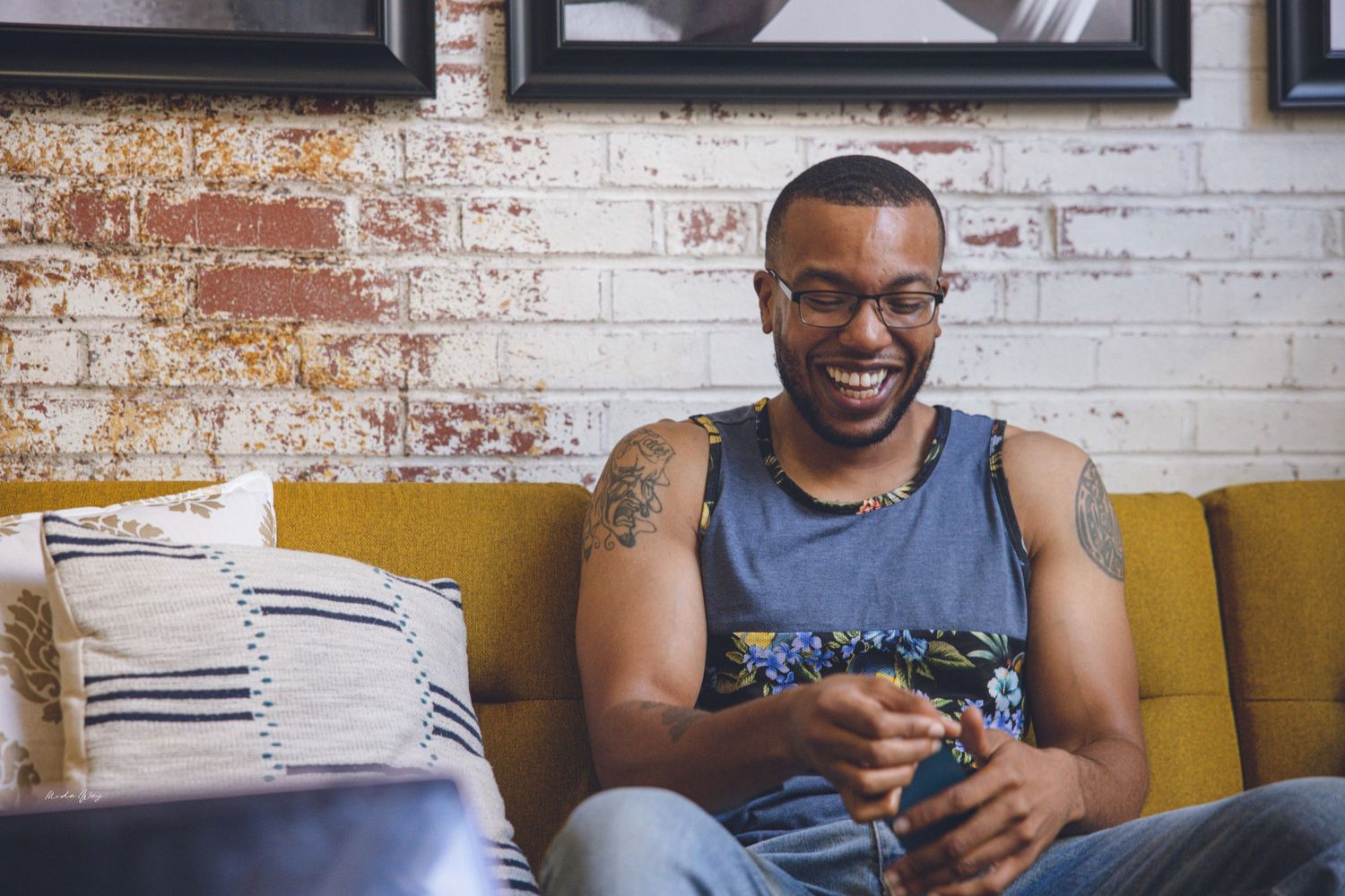 Person smiling while sitting on yellow couch with tattoos visible on arms.