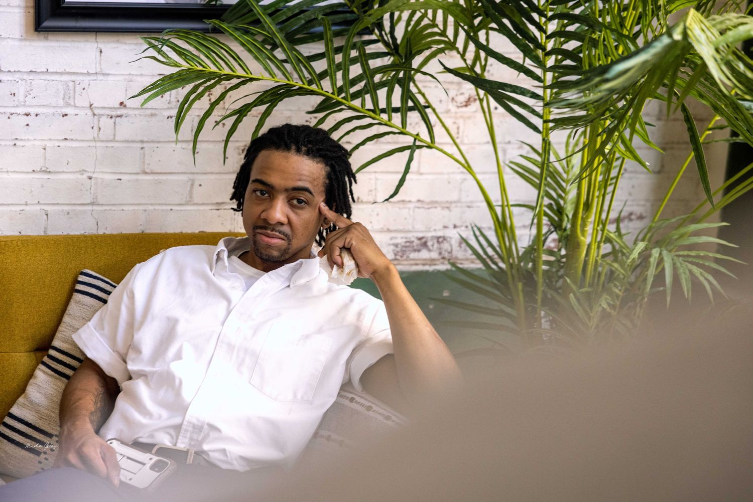 Person relaxing on yellow couch near indoor plants and white brick wall.