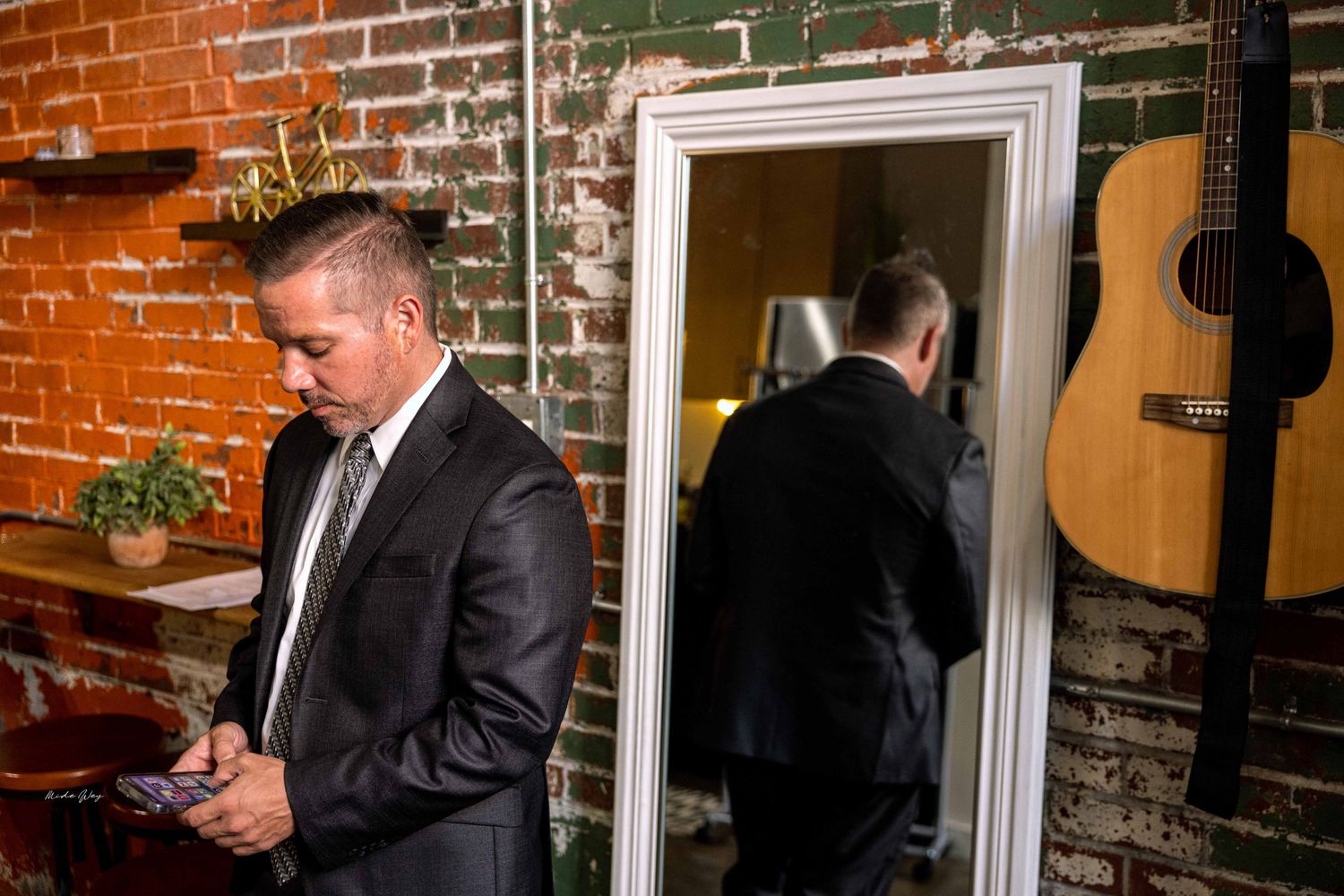Man in suit checks phone near mirror with acoustic guitar on brick wall.