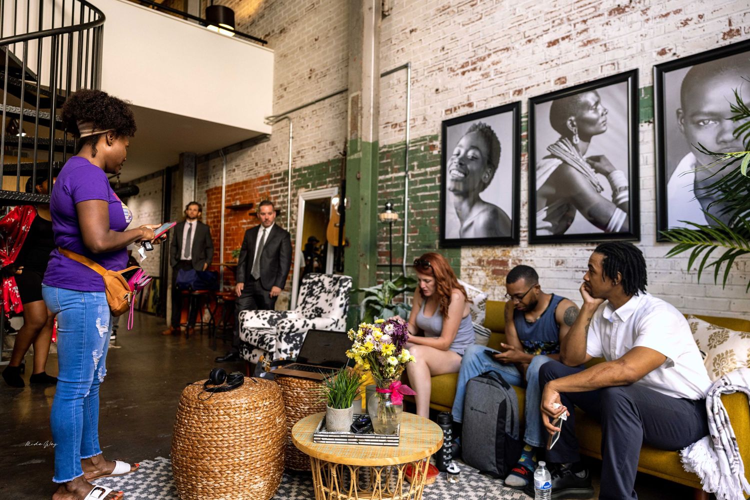 People gather in casual setting with vintage portraits on brick wall and wooden furniture.