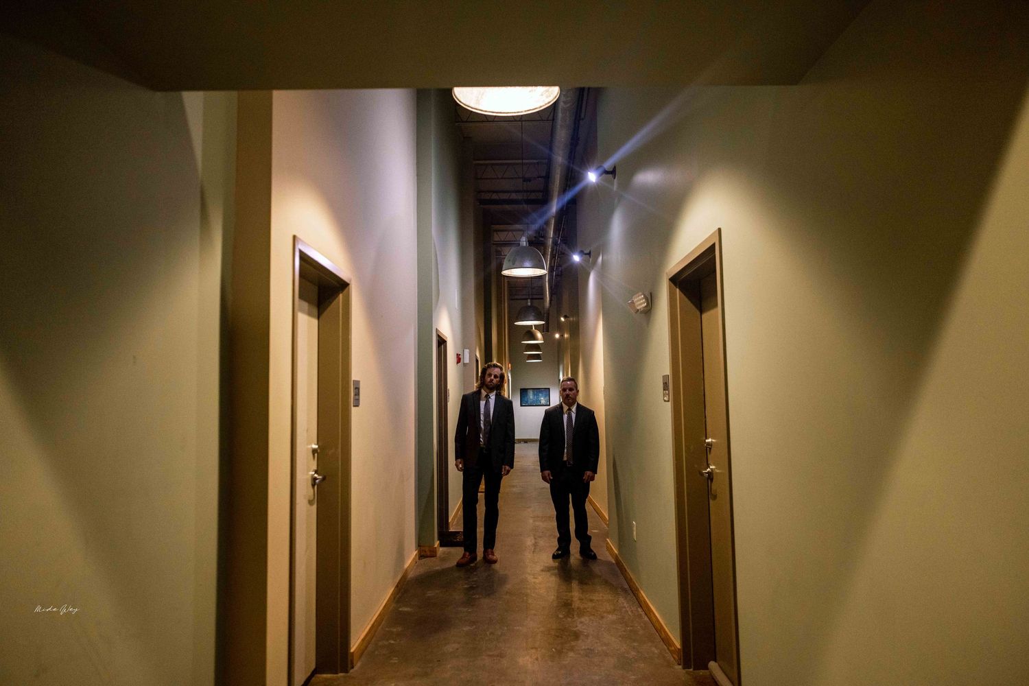 Two figures walk down a long, dimly lit corridor with doors on both sides.