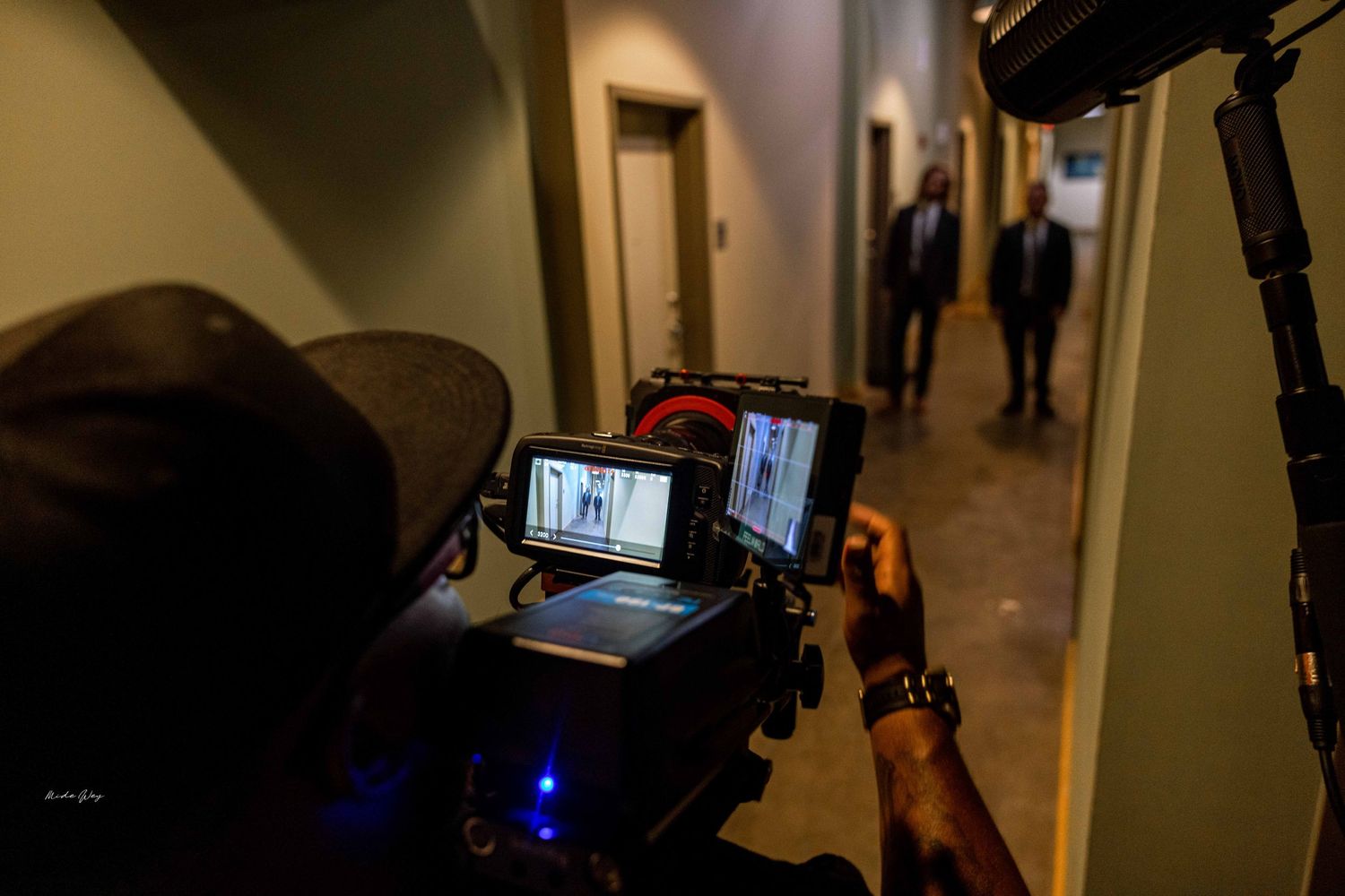 Camera operator films in hallway with people visible in background.