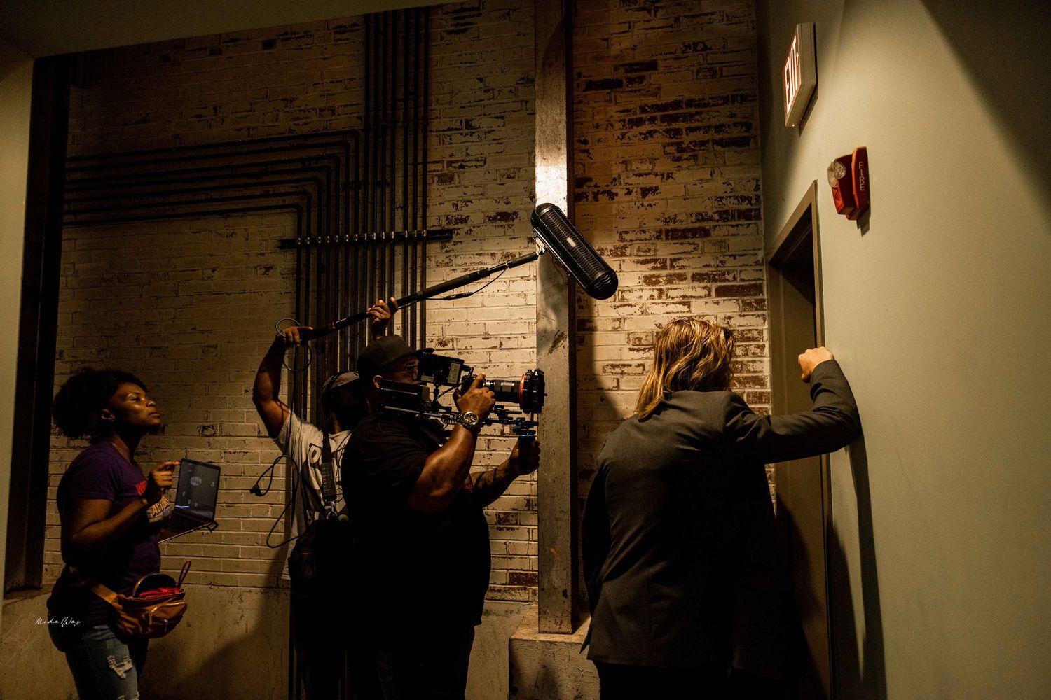 Film crew with camera equipment working against exposed brick wall.
