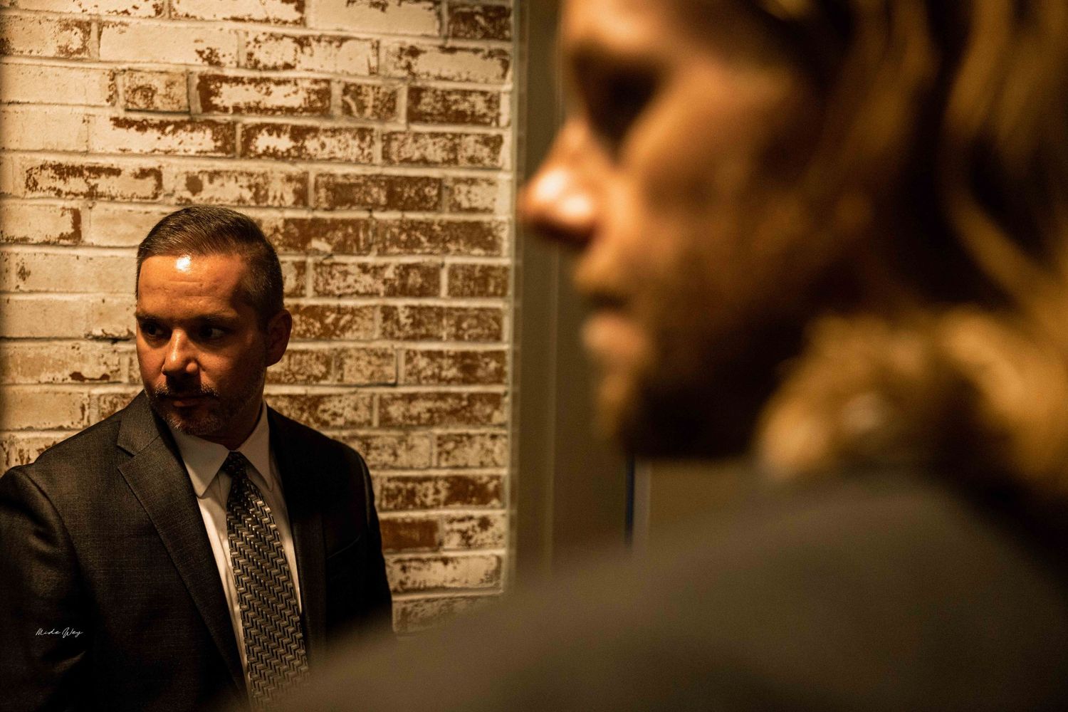A man in a dark suit stands against a textured brick wall in moody lighting.