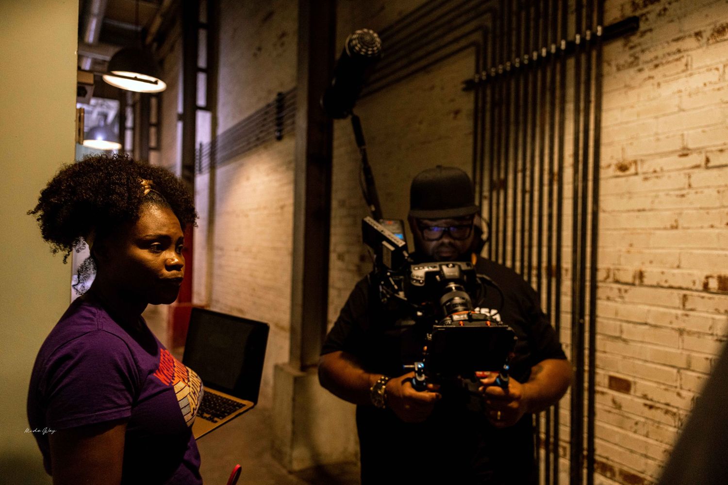 Production crew working with camera equipment in brick-walled hallway.