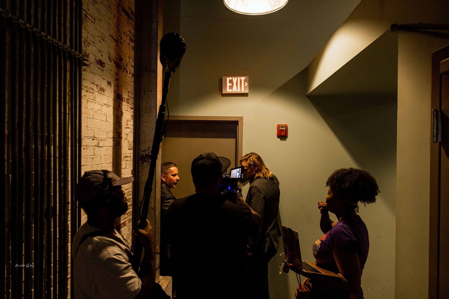 A group of people gather in a dimly lit hallway near an exit sign and brick wall.