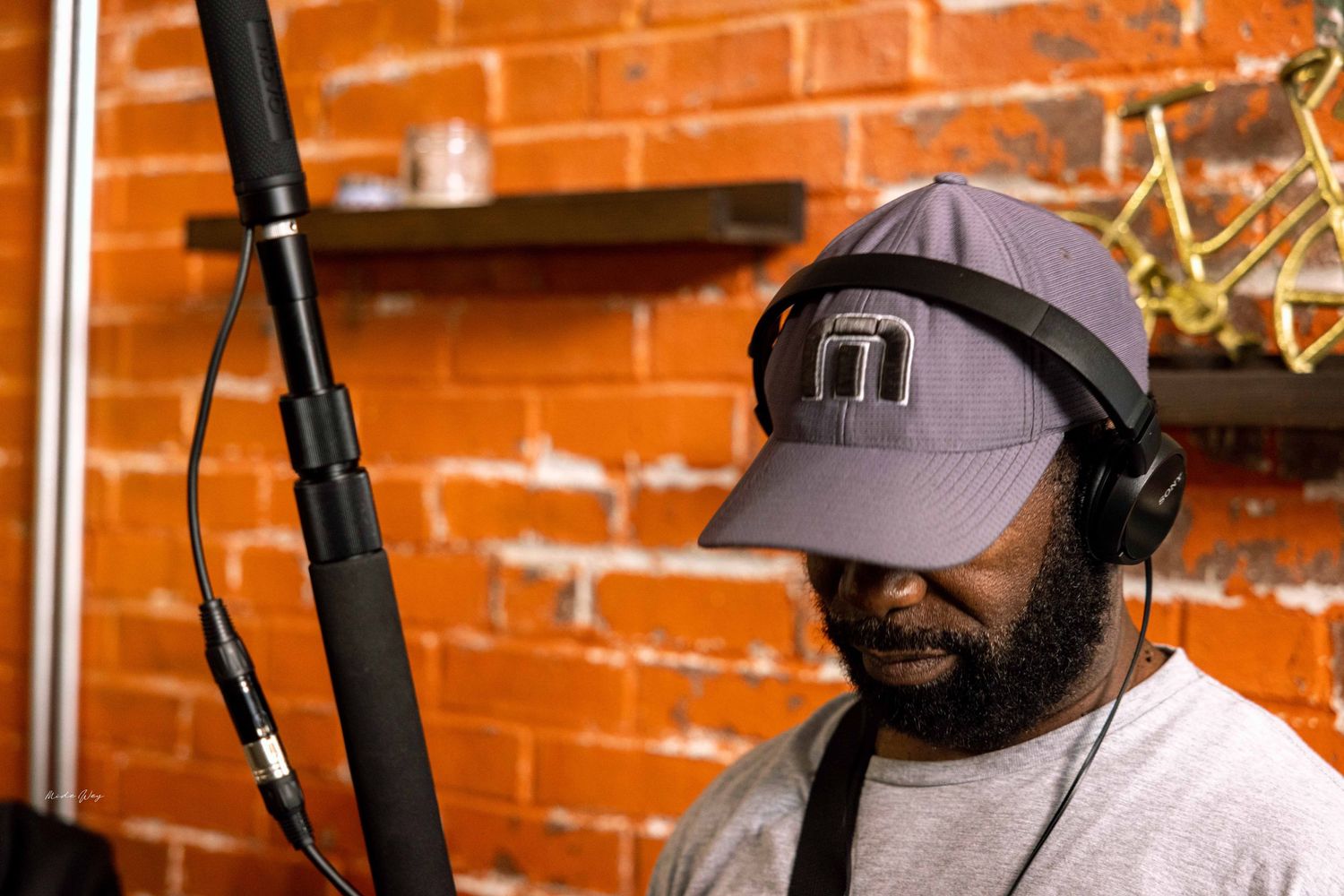 Person wearing headphones and cap speaking into microphone against brick wall.
