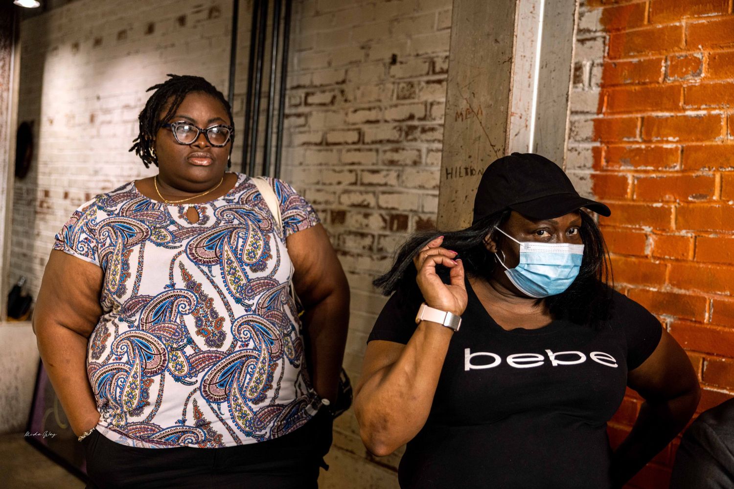 Two people seated against a brick wall, one wearing a face mask.
