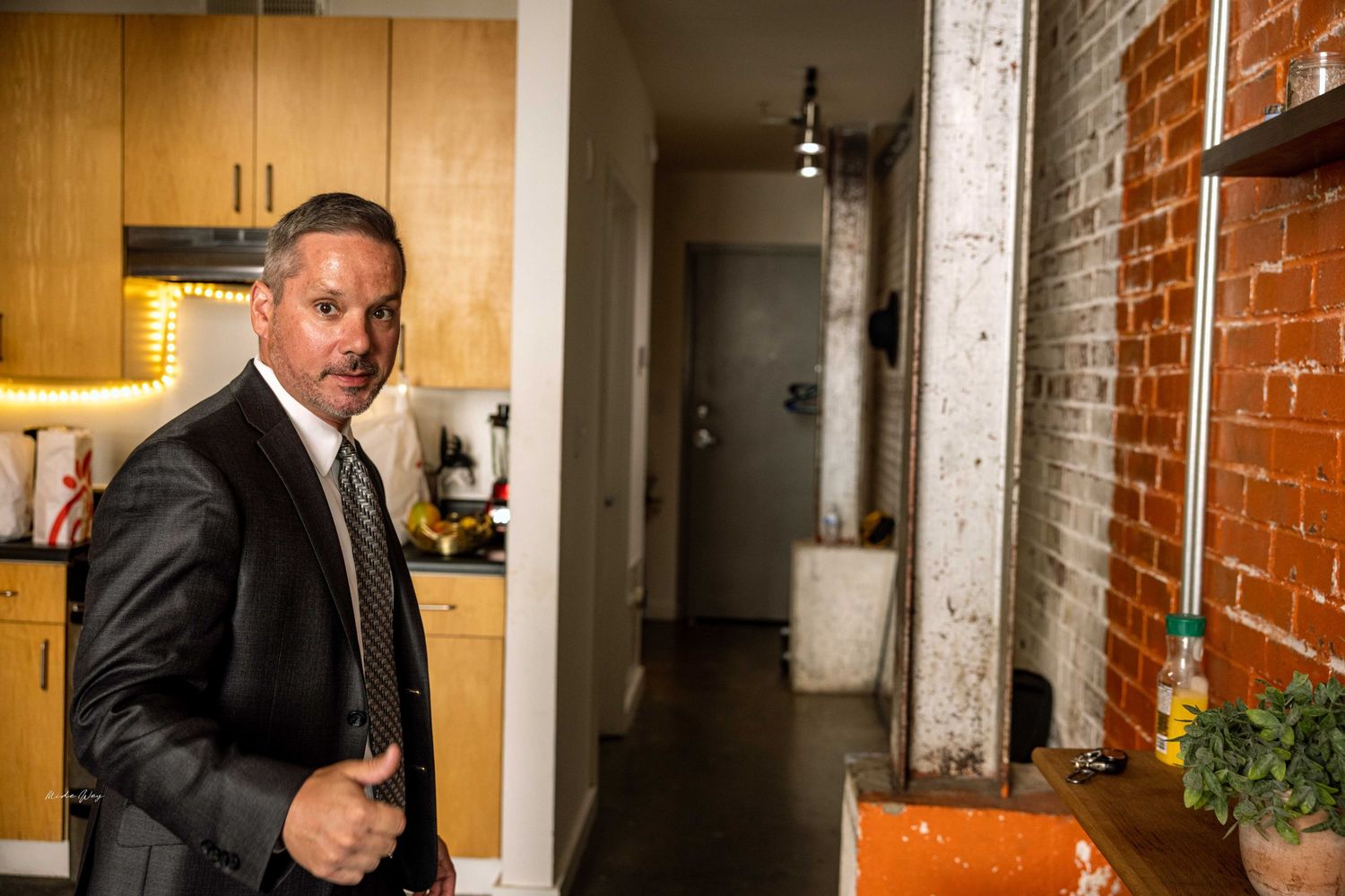 Professional in a business suit standing in a modern kitchen with exposed brick walls.