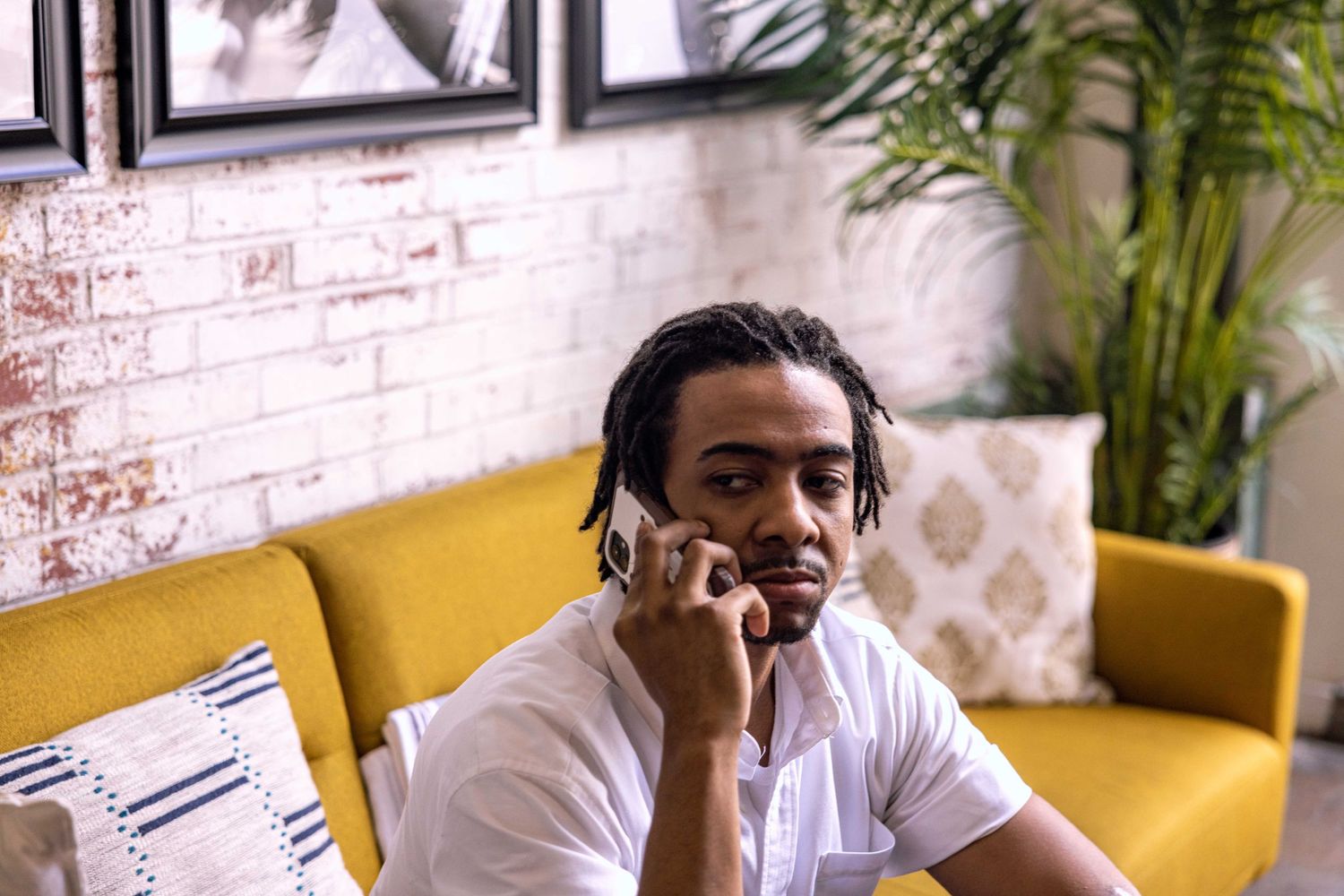 Person talks on phone while sitting on a yellow couch against exposed brick wall with plants.