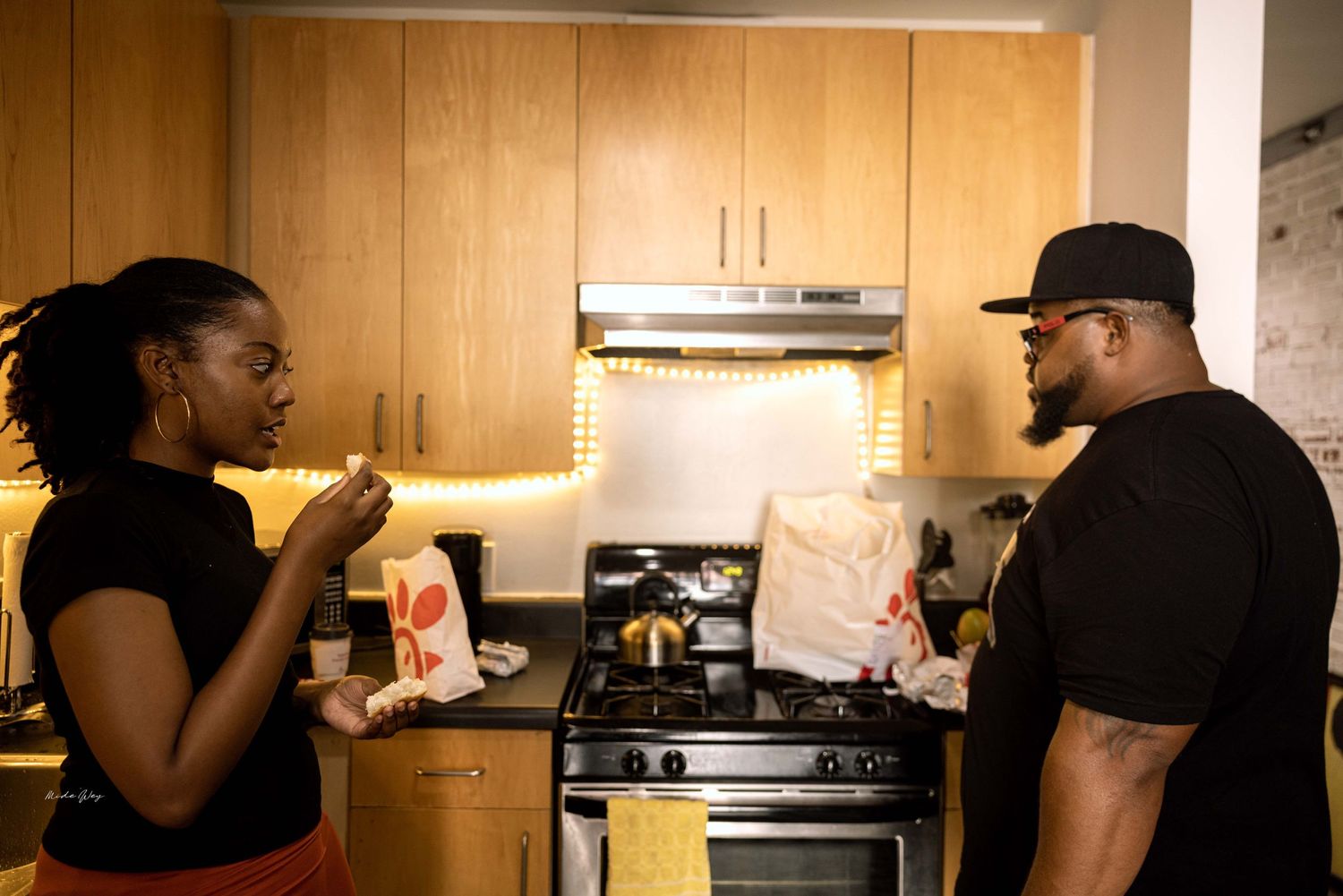 Two people having an animated conversation in a modern kitchen with wooden cabinets.