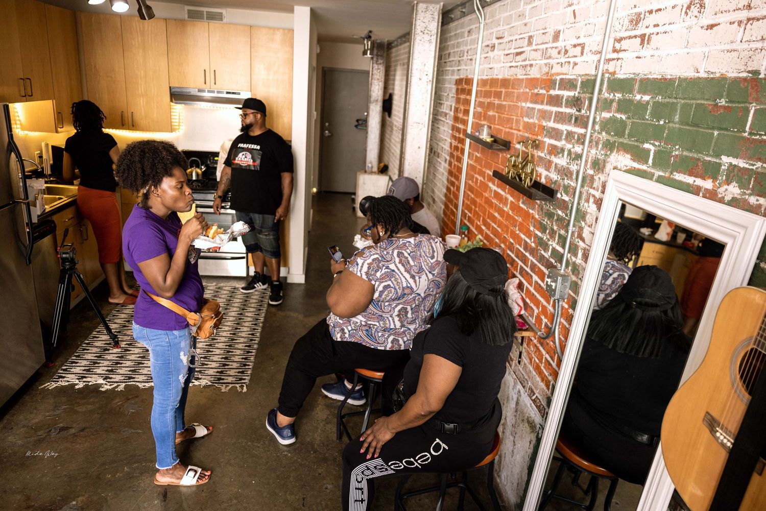 People gather in a casual indoor setting with exposed brick walls and kitchen area.