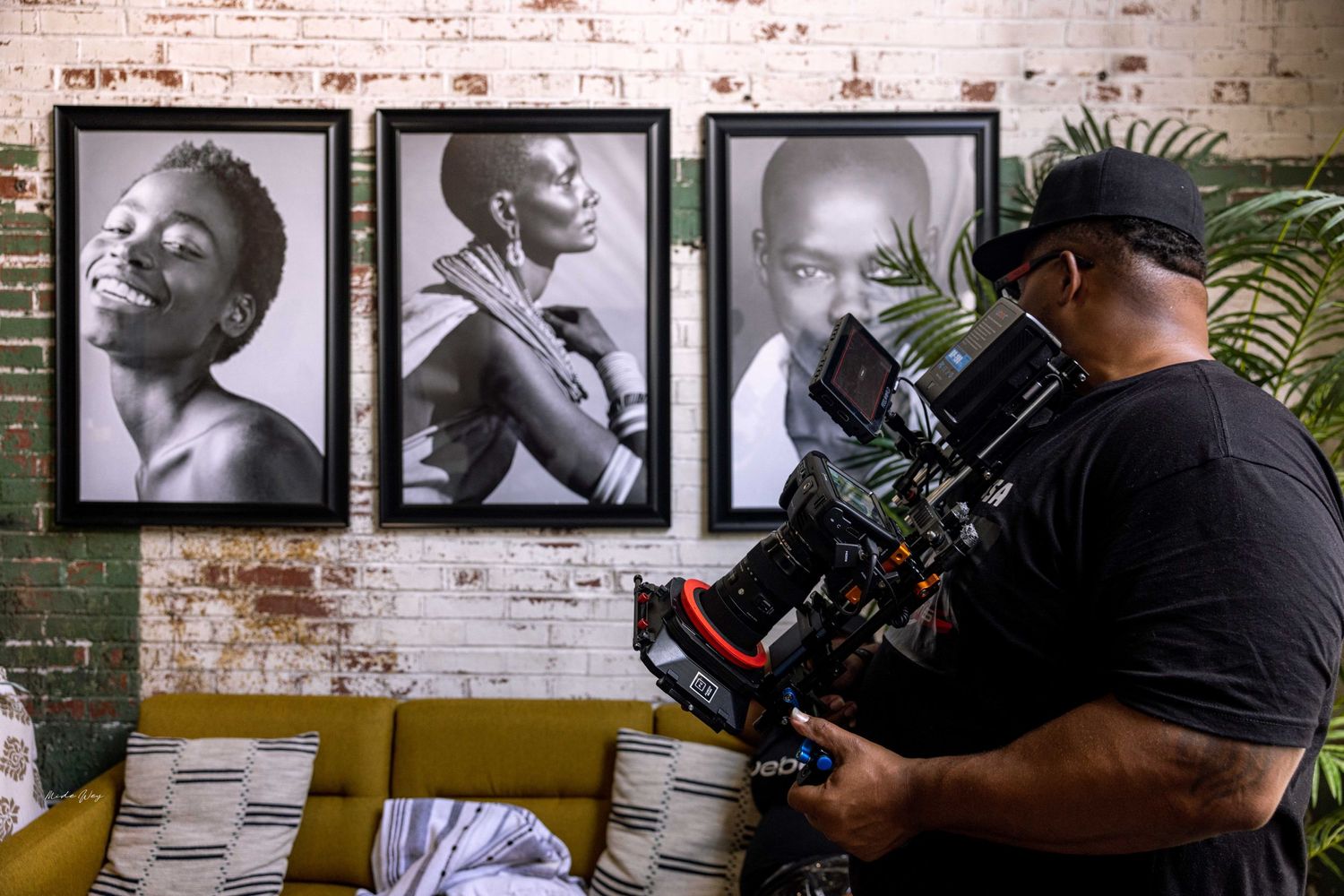 A camera operator films in front of framed black and white portraits on a brick wall.