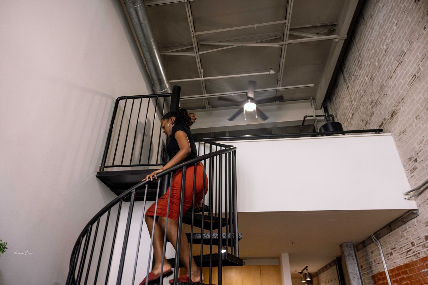 A person in a red dress descends a black spiral staircase in an industrial loft setting.