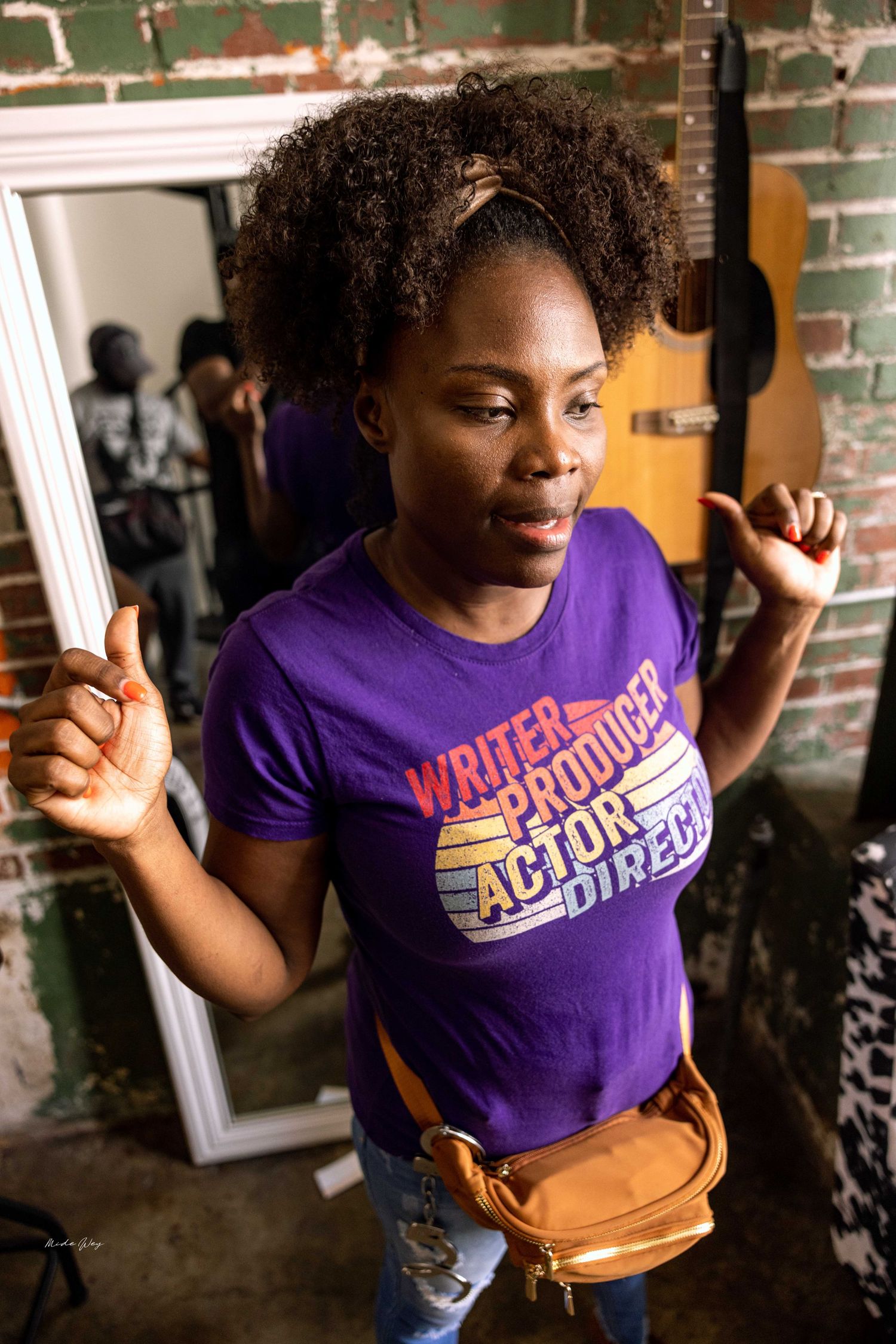 Person in purple t-shirt poses energetically against brick wall during video production.