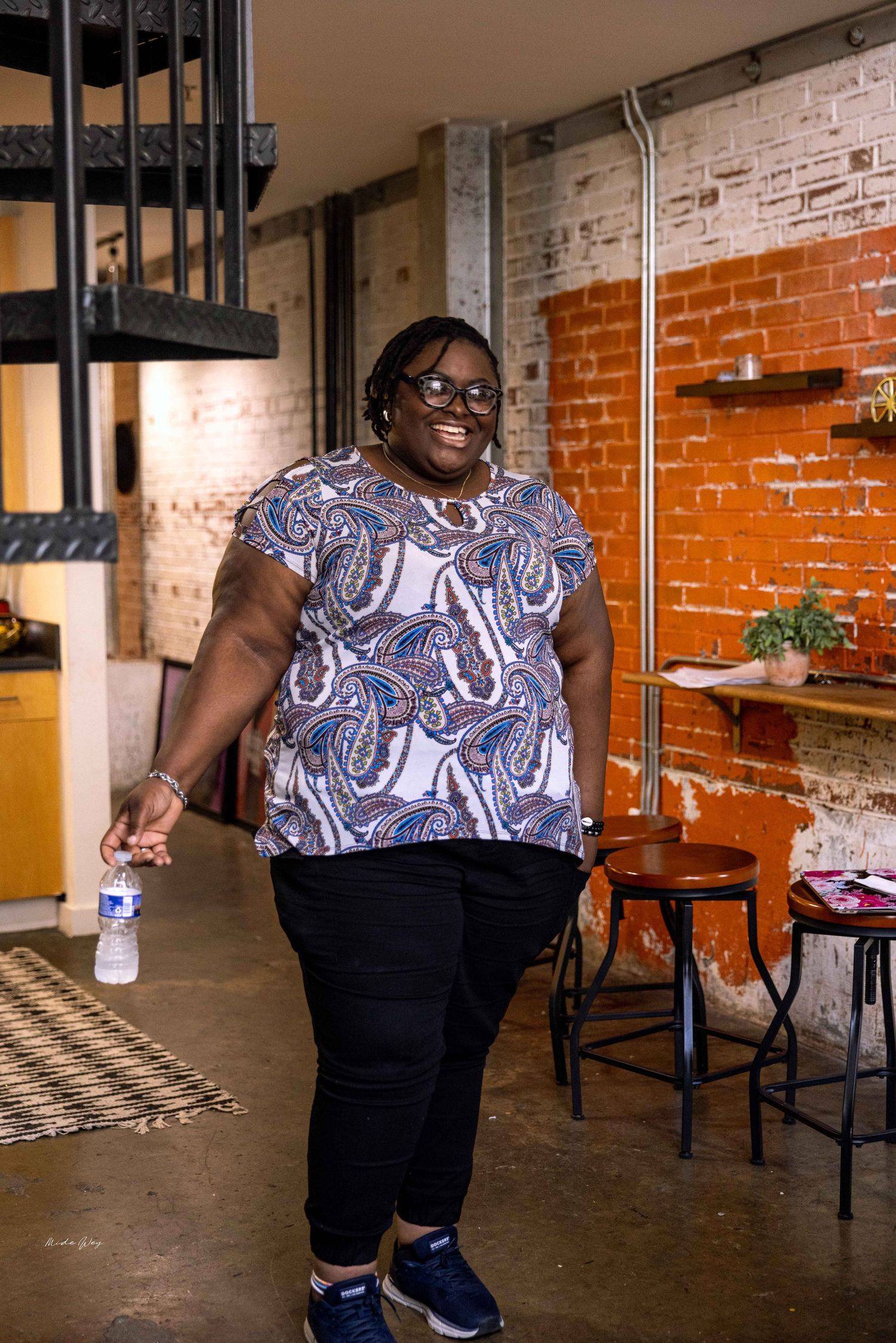 Person in patterned top and black pants poses playfully in industrial-style room with brick walls.
