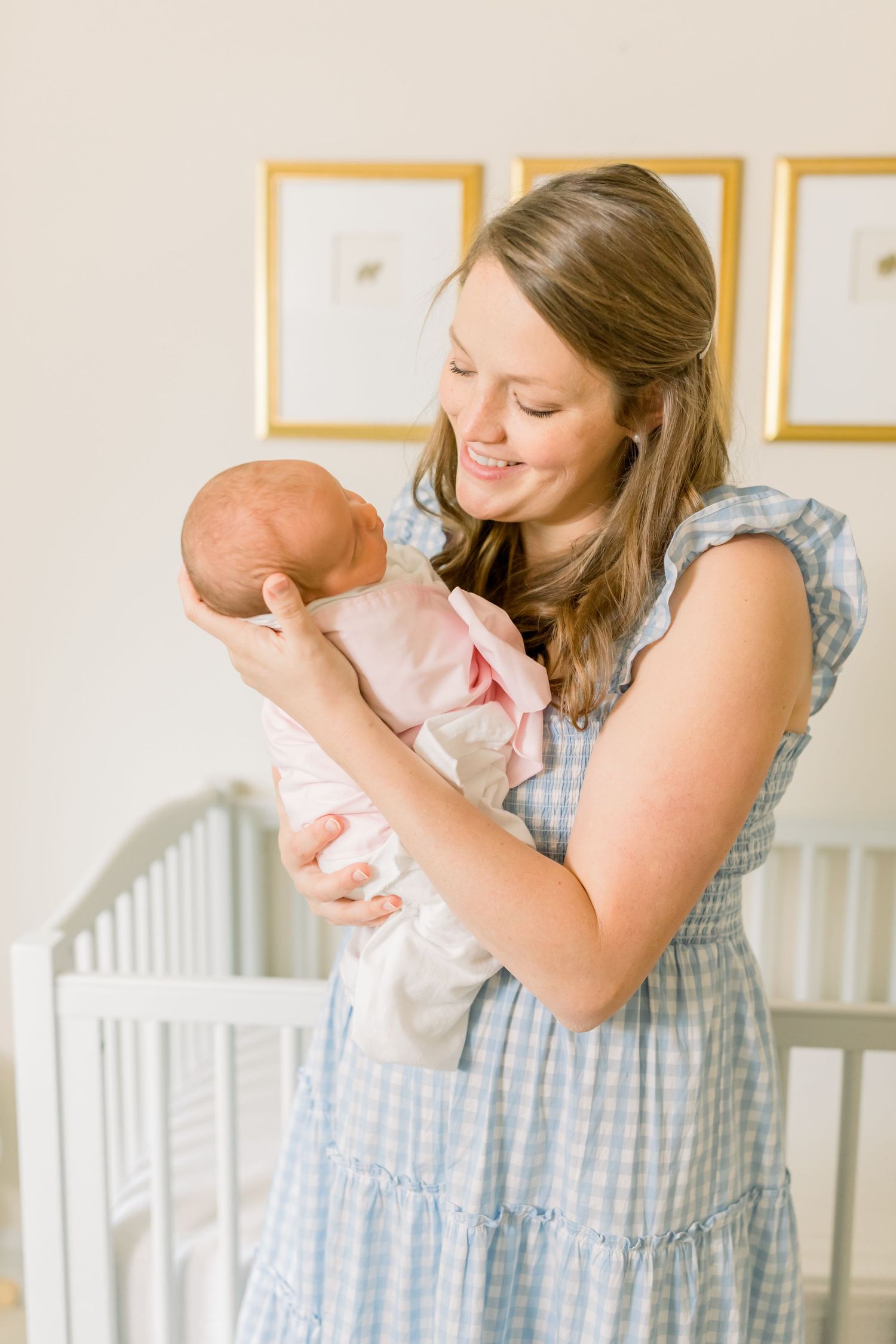 Mother in blue dress holds newborn baby near white crib with gold framed artwork on wall.