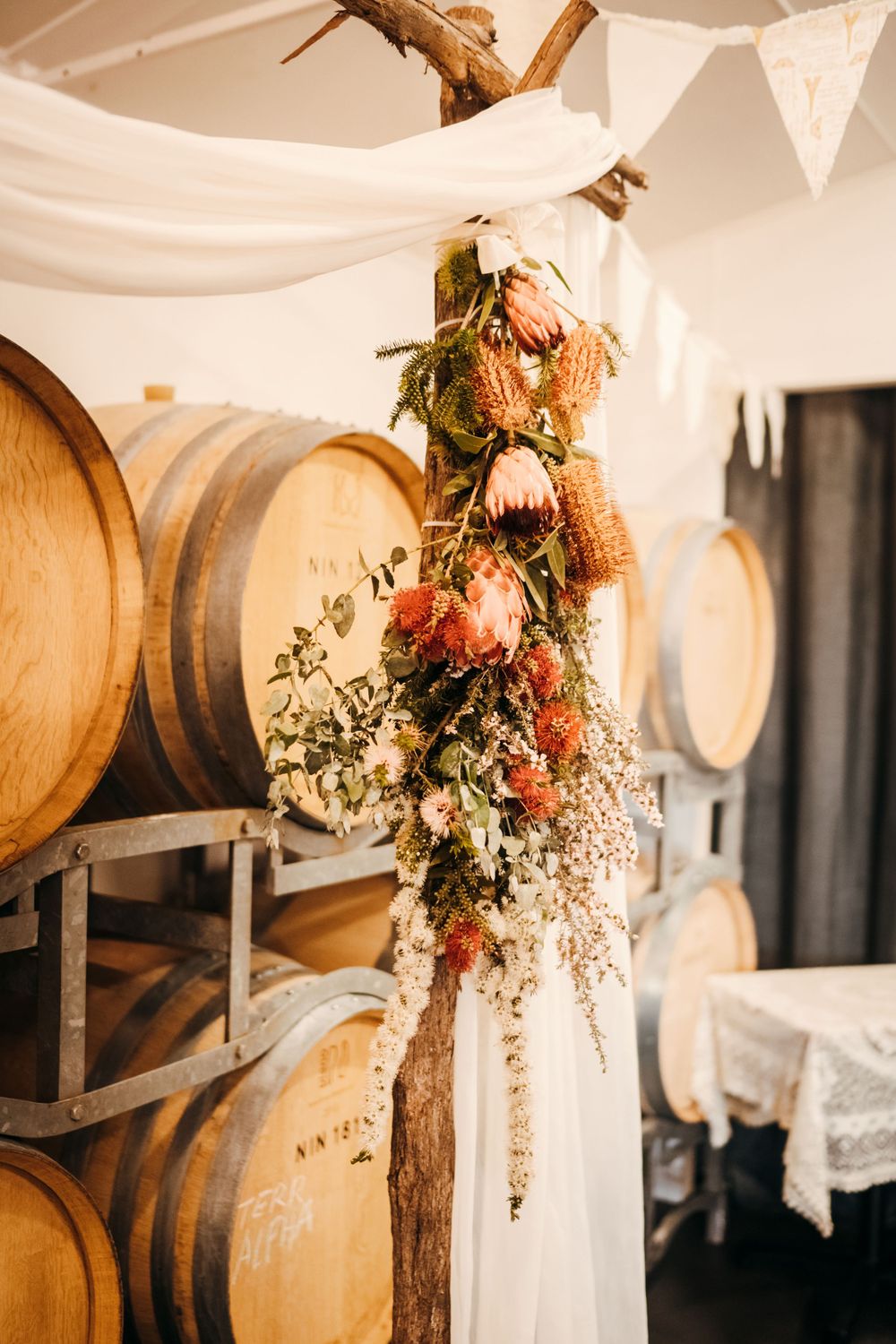 Dried floral arrangement hanging from wooden branch with wine barrels and white fabric draping in background.