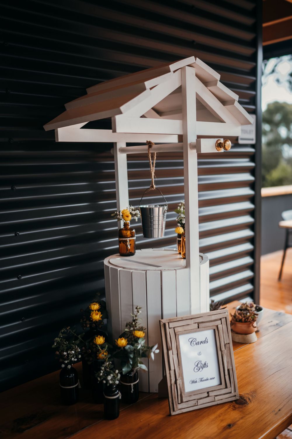 Decorative white wishing well display with flowers and a frame on a wooden table against a corrugated wall.