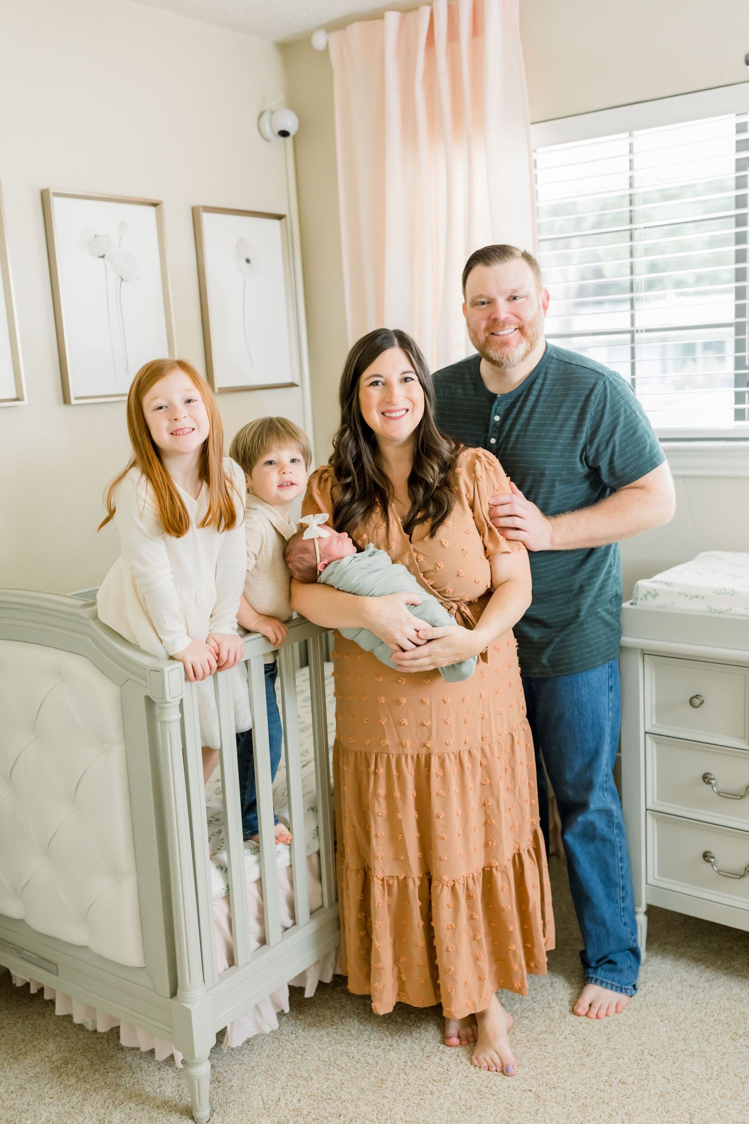 A family poses together in a nursery with gray furniture and peach curtains.