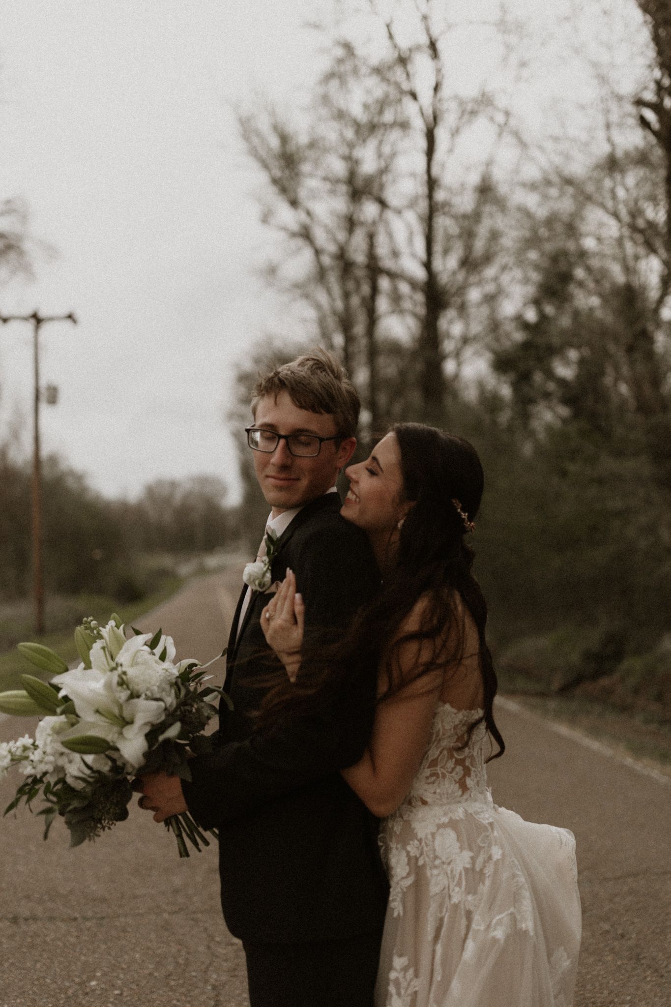 A joyful wedding couple sharing a laugh on a rural road with a white bouquet.