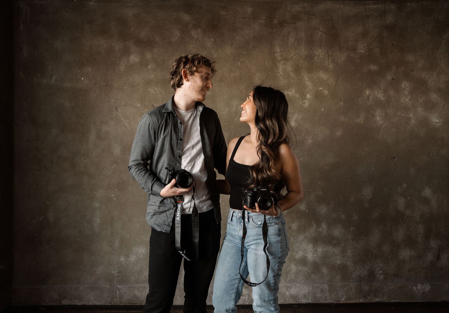 Young couple in casual attire having a candid moment against a textured gray wall.