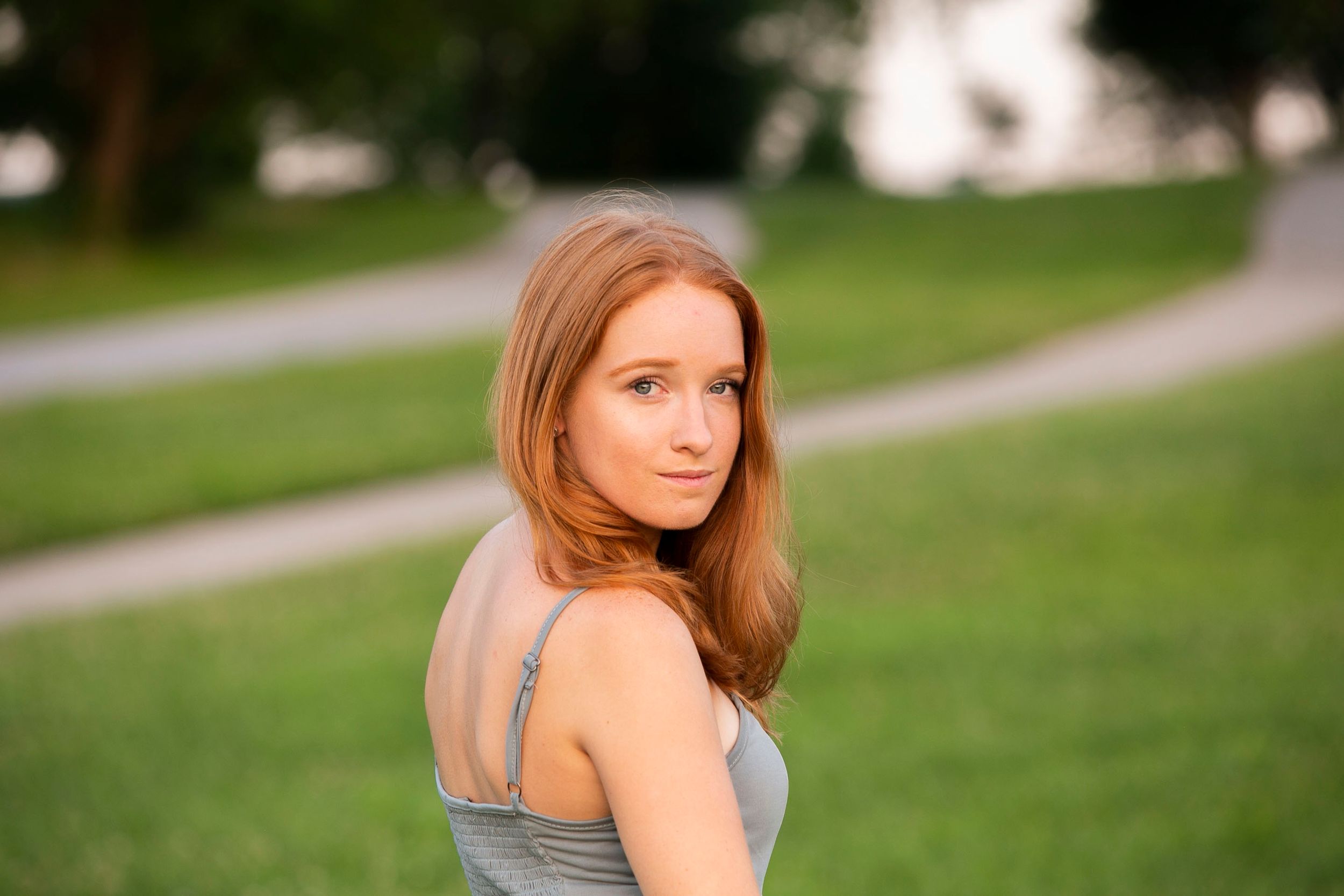 Young woman with red hair in a gray top posing outdoors in a green park.