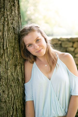 Young woman in light blue top leaning against a tree trunk outdoors.