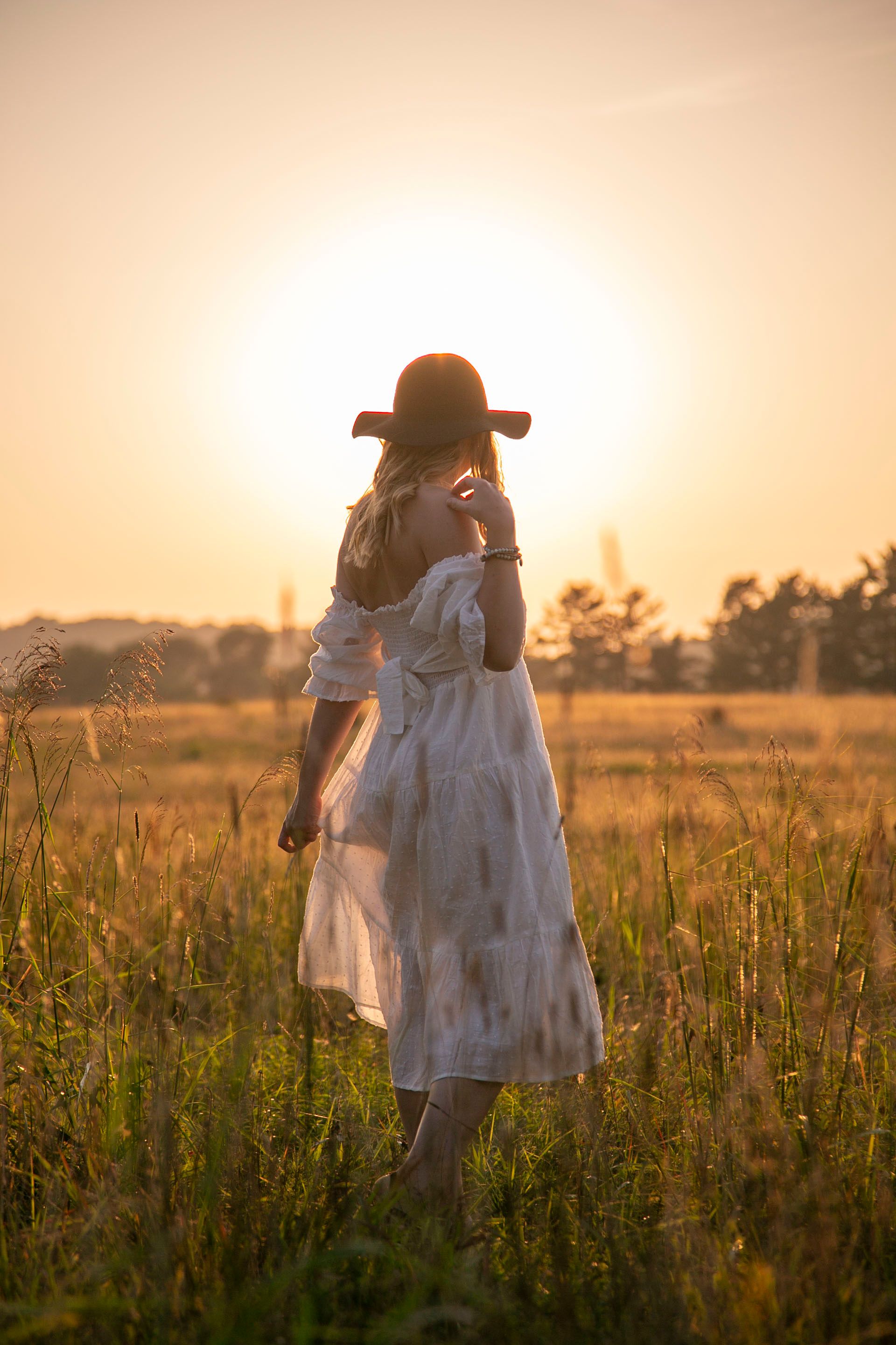 Woman in white dress and black hat standing in golden field at sunset.