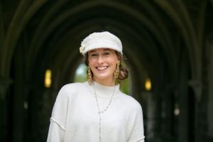 Smiling young woman in white sweater and beret standing under stone arches.