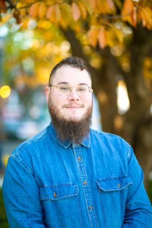 Bearded man in denim shirt smiling outdoors with autumn foliage background.